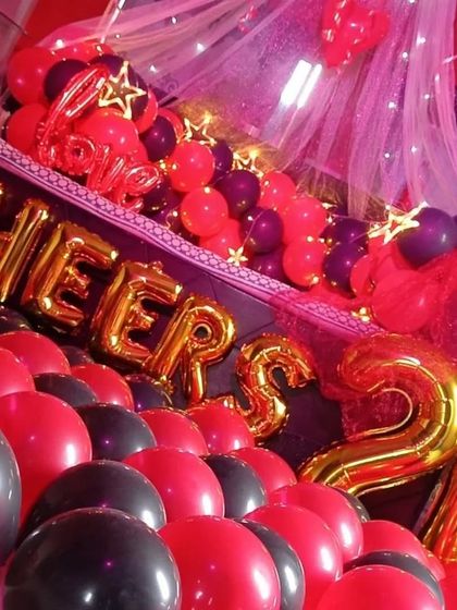 A close-up view of a 'Cheers to 20' celebration. This decoration uses red and black balloons, gold foil letters, and a romantic canopy with pink lighting to create a vibrant and celebratory mood.