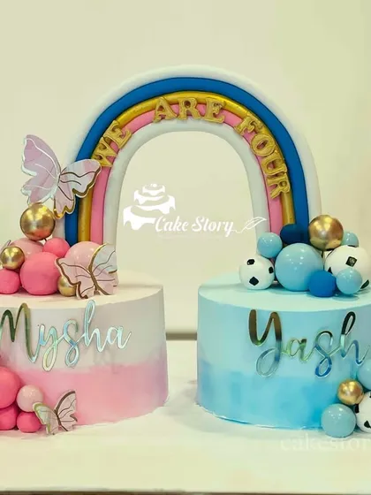 A creative cake set for twins or a baby shower. Two separate cakes, one pink for a girl and one blue for a boy, are connected by a rainbow that says 'We Are One'.
