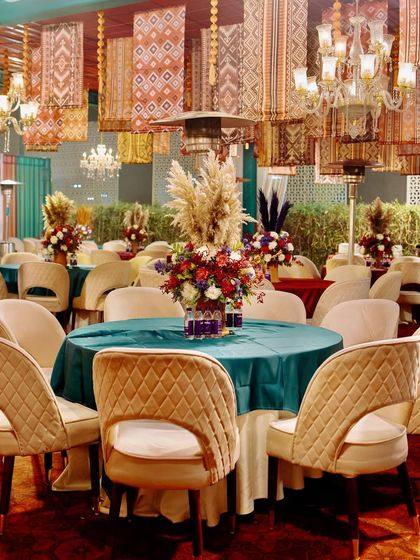 A close-up of a guest table, featuring a rich teal tablecloth, modern cream chairs, and a vibrant floral centerpiece with pampas grass.