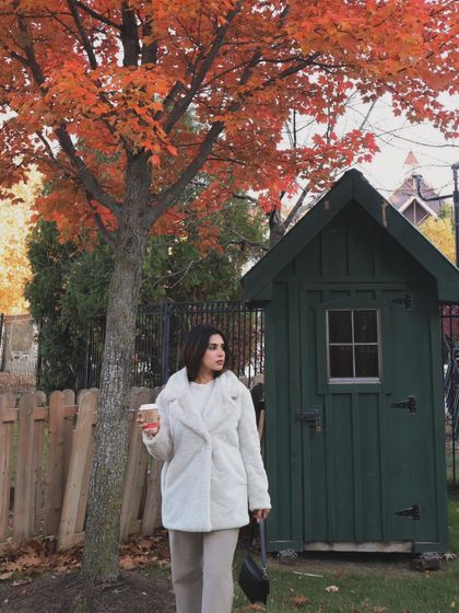 Posing with a coffee cup next to a charming green shed and a brilliant orange maple tree, perfectly capturing the fall vibe.