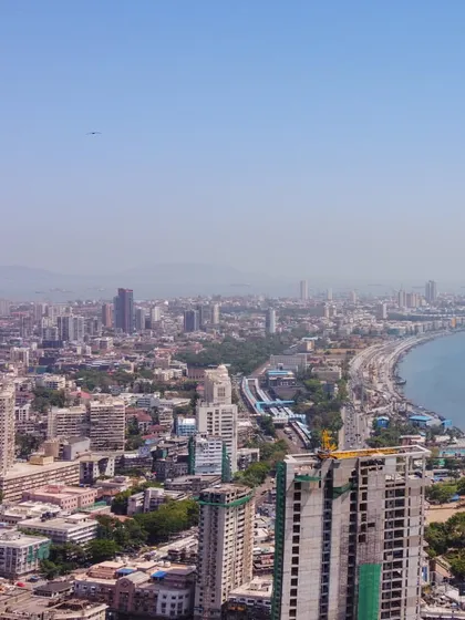 A daytime drone panorama showing the curve of Marine Drive in Mumbai, with the city on one side and the Arabian Sea on the other.