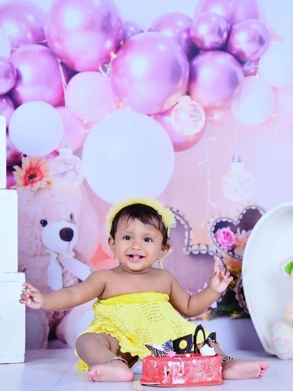 A first birthday cake smash scene with a teddy bear and balloons in the background.