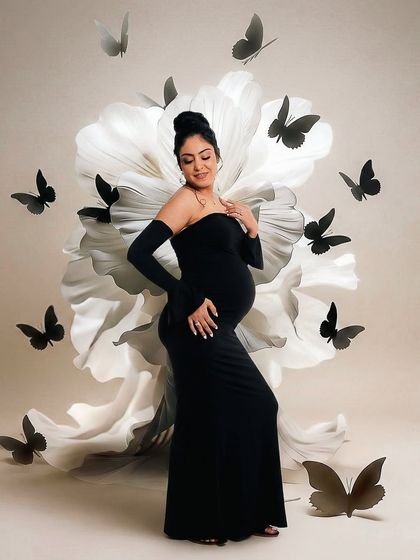A creative and artistic portrait featuring a mother-to-be in a black gown, surrounded by a large white paper flower and black butterfly cutouts. This setup adds a touch of fantasy and elegance.