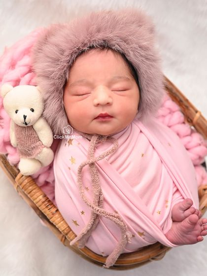 A cozy portrait of a baby girl in a pink furry hood, snuggled in a basket with her teddy bear. The pink tones create a warm and gentle feel.