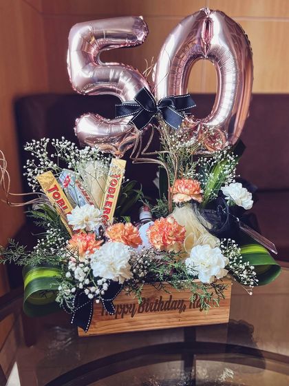 A 50th milestone birthday hamper designed for a refined taste. The composition balances celebratory balloons with sophisticated florals and gourmet chocolates in a personalized wooden crate.
