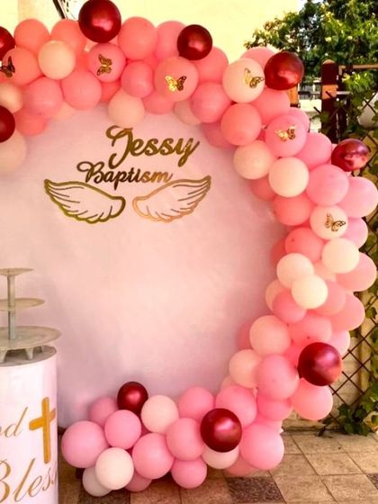 A pink-themed baptism decoration for a home setting. The circular backdrop is personalized and framed by a pink and chrome red balloon arch.