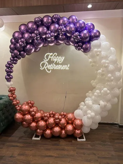 A sophisticated circular organic balloon arch in metallic purple, rose gold, and white for a retirement party. This modern design, complete with a "Happy Retirement" neon sign, honors a colleague's career in style.