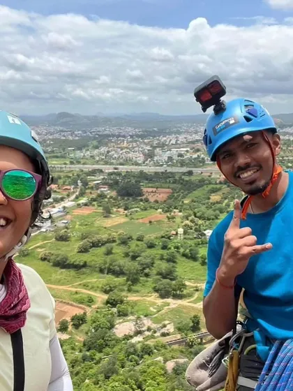 Akash and a friend share a smile and a shaka sign high up on a multi pitch route, with the city sprawling below.