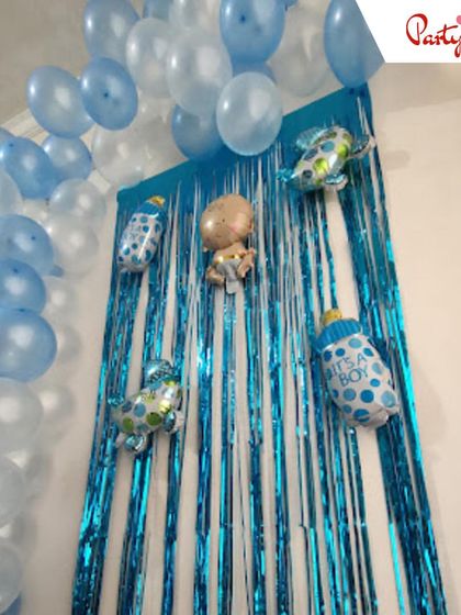 This "It's a Boy" welcome home decoration features a blue fringe curtain backdrop adorned with baby-themed foil balloons. A simple balloon arch in shades of blue and white frames the setup perfectly.
