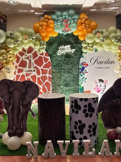A fun jungle animal theme for a first birthday party. This setup includes a green foliage backdrop, large animal cutouts of an elephant and gorilla, and a balloon arch in safari-inspired colors.