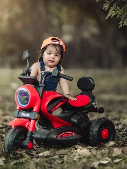 Your first breath took ours away. A toddler in a cool cap and overalls is ready to ride his red toy motorcycle.