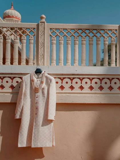 A detail shot of the groom's sherwani and necklace, hanging against a beautiful architectural backdrop in Jaipur, capturing the elegance of the wedding attire.