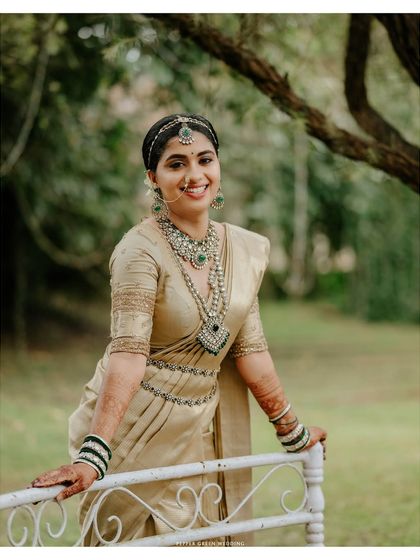 A joyful portrait of the bride in her golden wedding saree, her smile radiant and welcoming. The natural green background makes her traditional attire stand out beautifully.