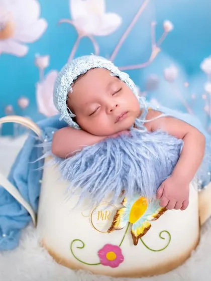 A sleeping newborn in a blue bonnet, nestled inside a decorative watering can prop against a dreamy blue floral background.