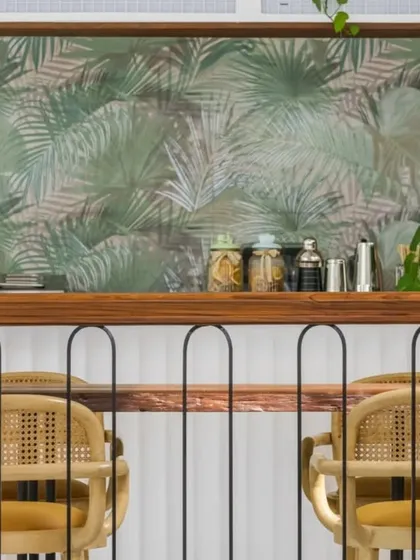 The bar area is highlighted by a beautiful tropical leaf wallpaper, adding depth and a focal point to the space. The use of natural wood for the bar top and rattan for the stools continues the cohesive, earthy material palette.