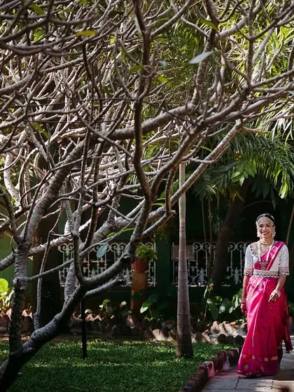 A bride in a bright pink saree walks through a grove of trees, a beautiful shot that highlights the natural beauty of the venue.