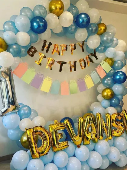 A first birthday decoration with a blue, white, and gold balloon arch, personalized with the name "Devansh" in foil letters.