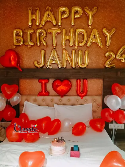 A romantic birthday decoration for your partner. This bedroom setup features red heart-shaped balloons, 'Happy Birthday Janu' and 'I Love You' foil letters, and a cake ready for celebration.