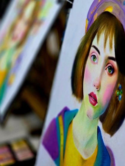 A close-up of a colorful portrait in progress, showing the rich texture and blending of soft pastels. The blurred background hints at a busy, creative classroom environment.