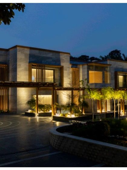The farmhouse exterior illuminated at night. The landscape lighting design highlights the architectural features, the circular driveway pattern, and the surrounding trees, creating a serene and dramatic ambiance.