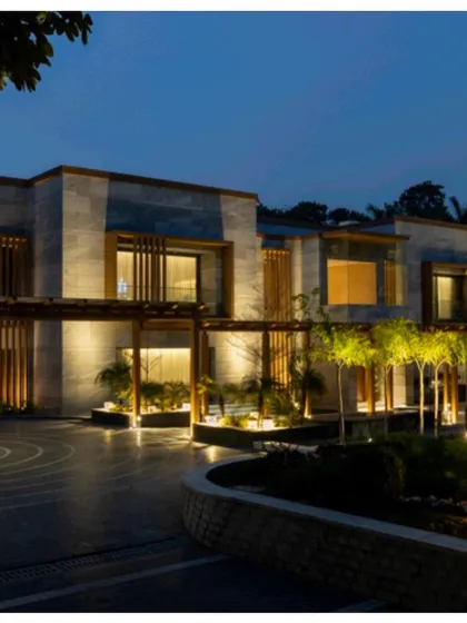 The farmhouse exterior illuminated at night. The landscape lighting design highlights the architectural features, the circular driveway pattern, and the surrounding trees, creating a serene and dramatic ambiance.