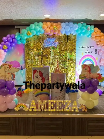 A large-scale unicorn theme on a stage. The rainbow balloon arch against a gold shimmer backdrop and multiple unicorn balloon columns make for a grand celebration.
