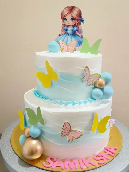 A two-tier butterfly themed cake with a soft blue and pink ombre effect and a cute doll topper.