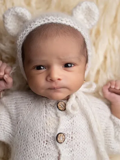This awake baby in a teddy bear hat is holding onto her parent's fingers, looking intently at the camera. A beautiful moment of connection.