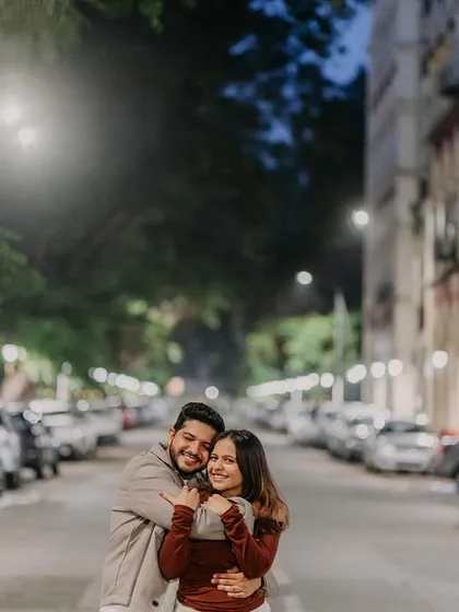 A cozy, candid hug on a quiet, rain-slicked street in Mumbai. The soft streetlights and classic architecture create a warm, romantic atmosphere for this pre-wedding moment.