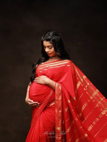 A quiet, contemplative portrait in a striking red saree. The focus is on the mother's gentle expression and the way she cradles the life within.