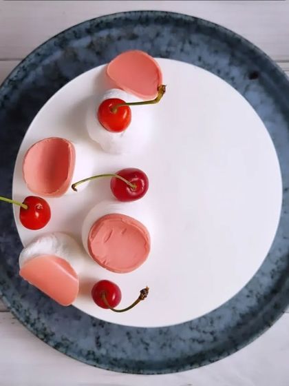 A minimalist and modern cherry mousse cake, with fresh cherries and delicate pink chocolate curls on a smooth white surface.