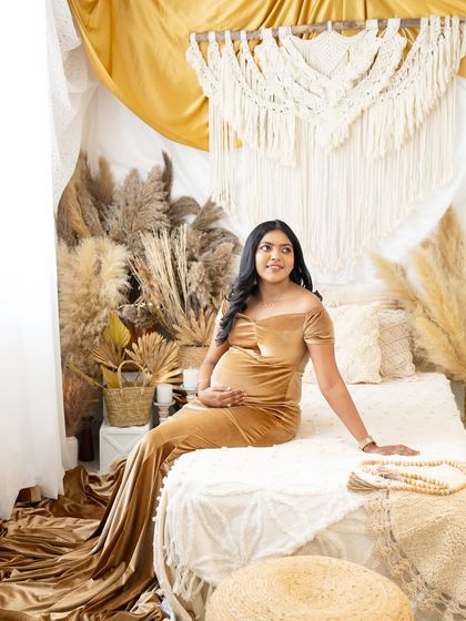 A solo portrait of a mother-to-be seated on a bed in a boho-styled room. She is wearing a beautiful golden velvet gown, looking towards the window with a happy expression.