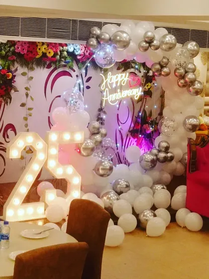 A 25th anniversary party decoration in a banquet hall. The setup includes a silver and white balloon arch, a neon sign, and large light-up numbers.