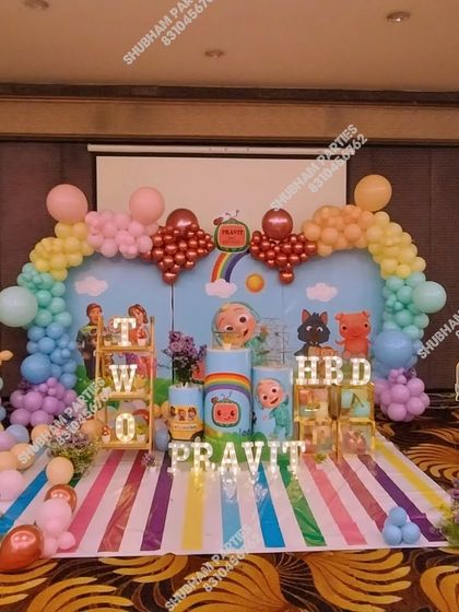 An elaborate Cocomelon stage decoration for a second birthday, complete with a rainbow balloon arch, character props, and a striped floor mat.
