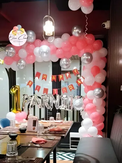 A pink, white, and silver balloon arch set up inside a restaurant for a first birthday.