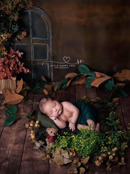 A cute little prince in his rustic home. The wooden backdrop, scattered leaves, and simple green pants create a charming and naturalistic scene.