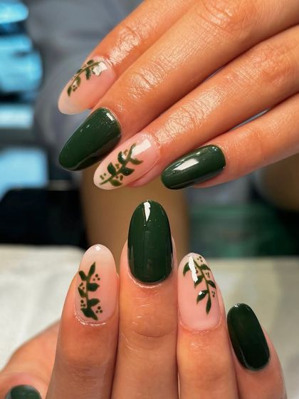 Green is the new black. This elegant manicure combines solid dark green nails with accent nails featuring delicate, hand-painted leafy branches on a sheer nude base.