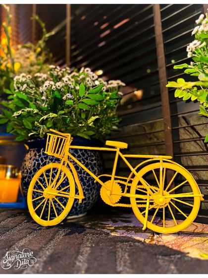 A whimsical detail, this miniature yellow bicycle prop adds a touch of playful charm to the decor, nestled amongst potted plants and candles.