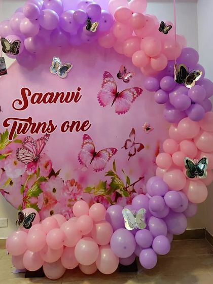 A beautiful butterfly-themed first birthday for "Saanvi." The decoration features a large pink circular backdrop with custom text and graphics, framed by a pink and purple balloon garland with butterfly accents.