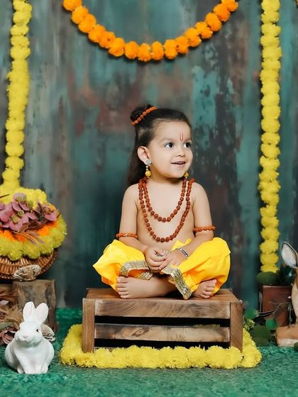 A serene moment, with 'Little Ram' sitting peacefully in a meditative pose amidst a rustic, forest-like setting with marigold garlands.