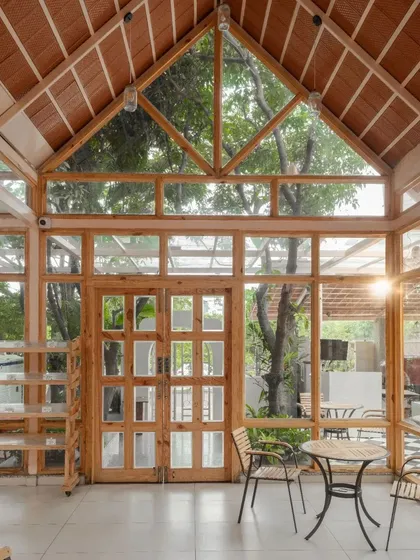 The main dining hall features a high, pitched roof and glass walls, creating a light-filled, greenhouse-like atmosphere. The wooden structure and large windows frame views of the surrounding trees.