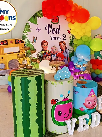 A complete Cocomelon-themed second birthday setup. This vibrant decor includes a rainbow balloon arch, the iconic yellow school bus, character-printed plinths, and a large light-up number '2'.