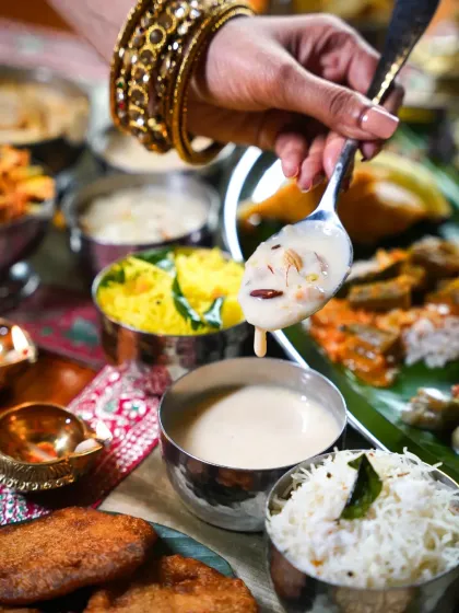 A hand with traditional bangles serving a spoonful of creamy payasam during an Onam feast. This action shot adds a human touch and a sense of celebration to the festive spread.