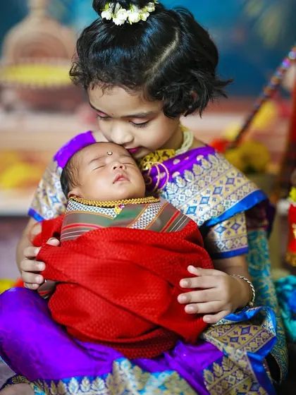 An older sister lovingly holds her newborn sibling, both dressed in beautiful traditional Paithani-style outfits for a festive family photo.