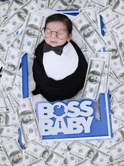 The ultimate Boss Baby. This wide shot shows the newborn sleeping peacefully on a bed of money, wearing a tiny tuxedo wrap and glasses. It's a fun and unforgettable theme.