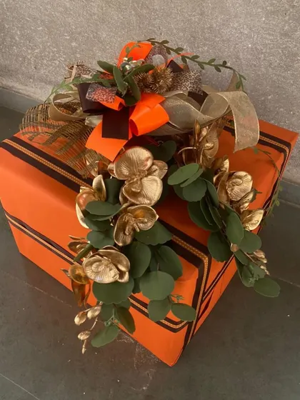 An exquisitely wrapped gift box in a vibrant orange, adorned with golden foliage and a multi-layered bow. This showcases our skill in creating standout packaging that makes the gift inside even more special.