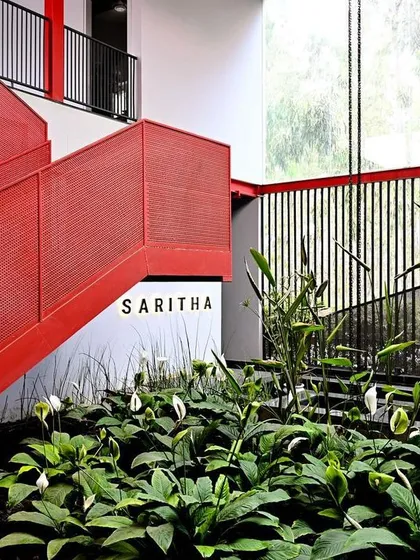 A clear view of the SARITHA brand signage, integrated into a clean white wall. The surrounding red architectural elements and lush greenery frame the entrance, creating a strong and cohesive brand environment.