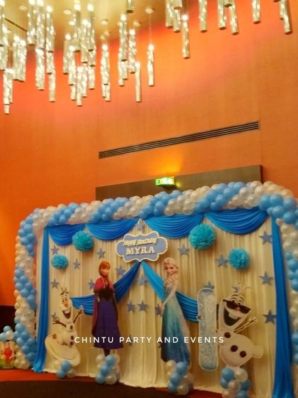 A Frozen themed first birthday party with a large stage setup. The blue and white decor includes character cutouts and a balloon arch over draped curtains.