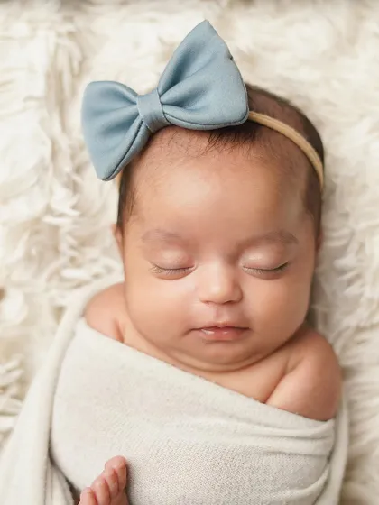 A close-up of a newborn girl with a sweet blue bow, sleeping soundly. The simple swaddle and soft background keep the focus on her adorable, peaceful expression.