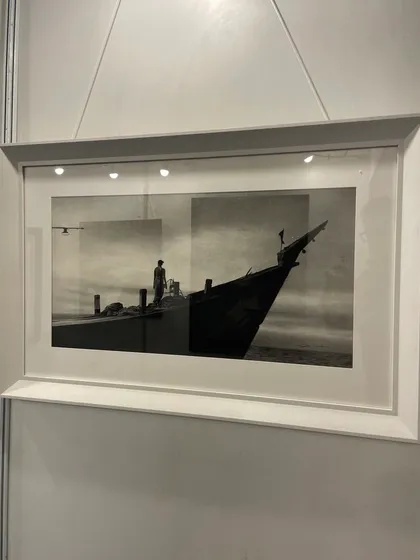 'Serene', an 18 by 36 inch charcoal drawing on white paper, shown here framed at an exhibition. The artwork captures a tranquil moment on the water.